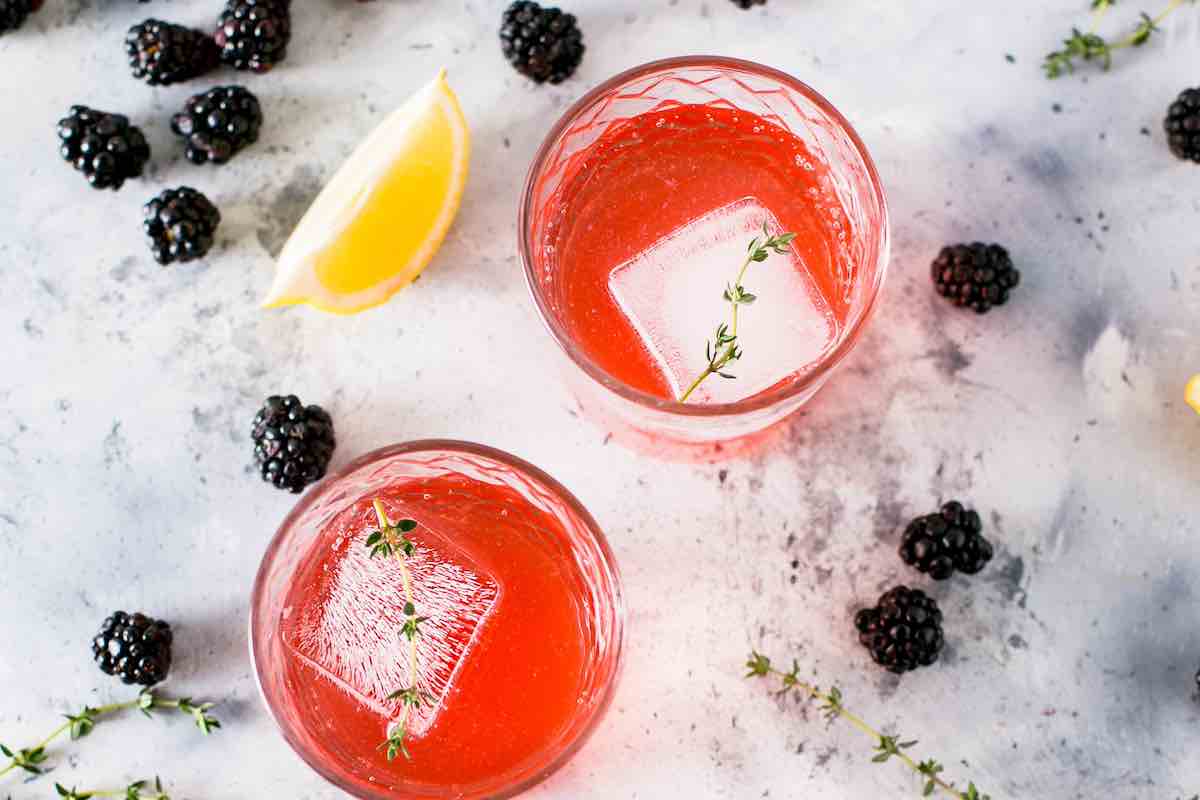 two red cocktails with large ice cubs, surrounded by blackberries and a lemon wedge
